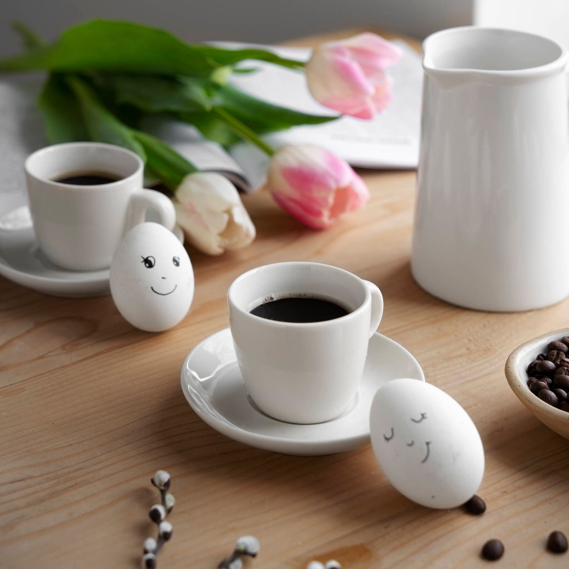 White coffee cups on wooden table with pink tulips and two hard boiled eggs with funny faces drawn on them