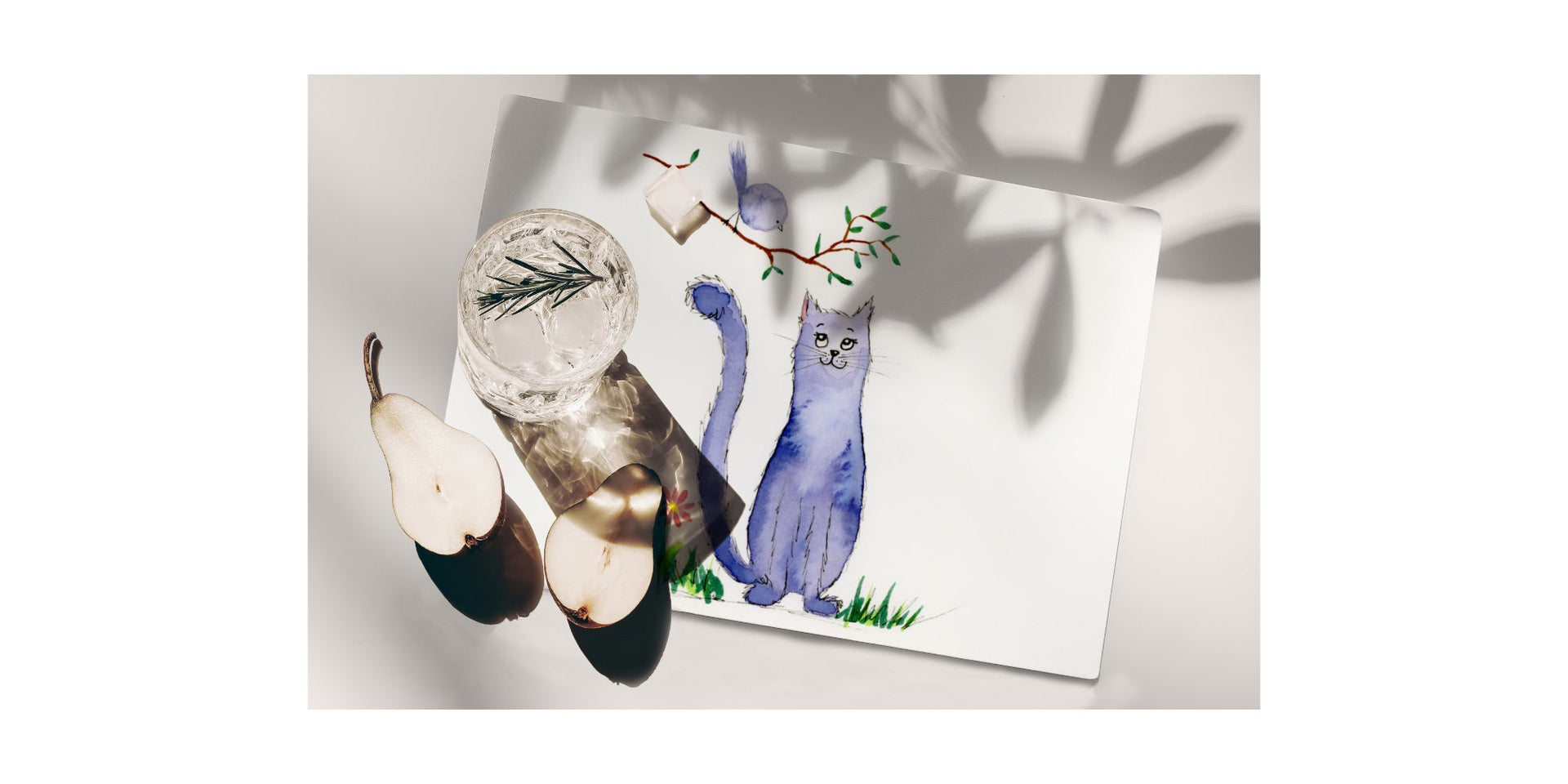 Rectangular placemat featuring a whimsical cat and bird artwork on a white table with a glass of water, icecube and sliced pear
