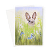 A vertical greeting card featuring a painted bunny in a grassy field with white flowers and bluebells, shown with a white envelope behind it on a light gray background.
