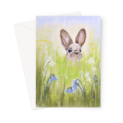 A vertical greeting card featuring a painted bunny in a grassy field with white flowers and bluebells, shown with a white envelope behind it on a light gray background.
