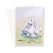 A greeting card featuring a soft pastel illustration of a fluffy white bunny holding a small blue watering can in a grassy field with white flowers, shown with a white envelope behind it on a light gray background.