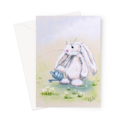 A greeting card featuring a soft pastel illustration of a fluffy white bunny holding a small blue watering can in a grassy field with white flowers, shown with a white envelope behind it on a light gray background.