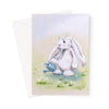 A greeting card featuring a soft pastel illustration of a fluffy white bunny holding a small blue watering can in a grassy field with white flowers, shown with a white envelope behind it on a light gray background.