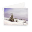 A greeting card featuring a painted winter landscape with snow-covered ground, evergreen trees, and a soft lavender sky, shown with a white envelope behind it on a light gray background.