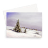 A greeting card featuring a painted winter landscape with snow-covered ground, evergreen trees, and a soft lavender sky, shown with a white envelope behind it on a light gray background.