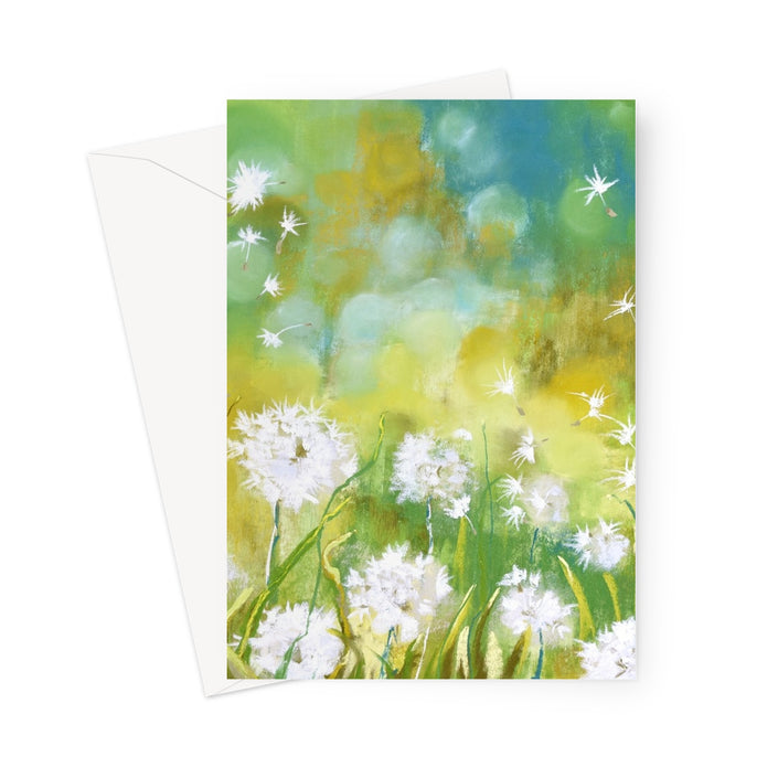 A vertical greeting card featuring a painted field of white dandelions with a soft green, yellow, and blue background, shown with a white envelope behind it on a light gray studio background.