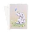 A greeting card with a white envelope behind it, featuring a soft pastel illustration of a fluffy white bunny holding small white flowers under a blue butterfly on a soft beige background.
