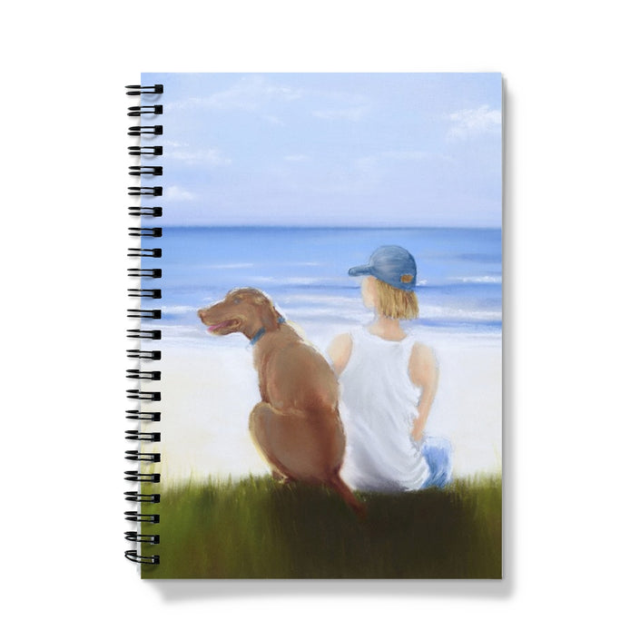 Notebook with a design of a boy and dog sitting on grass by the beach