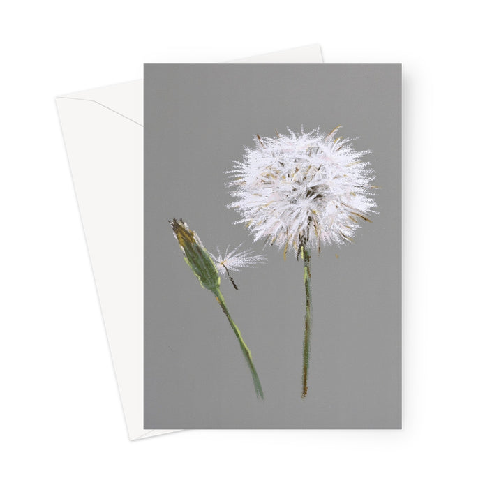 A greeting card with a dandelion seed head and bud on a grey background, shown with a white envelope behind it on a white studio background.