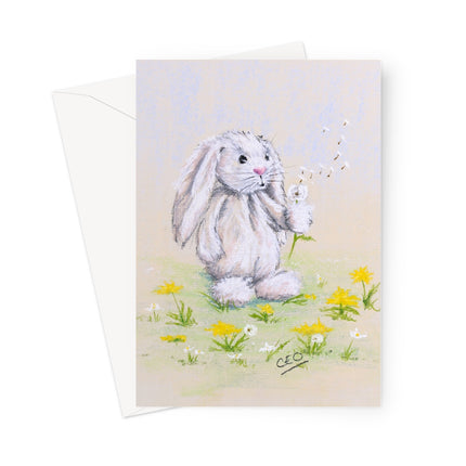 A rectangular art print or card featuring a soft pastel illustration of a white rabbit sitting in a field of yellow and white flowers, blowing dandelions. A white envelope is partially visible behind the card on a clean white background.