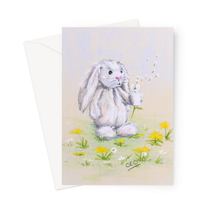 A rectangular art print or card featuring a soft pastel illustration of a white rabbit sitting in a field of yellow and white flowers, blowing dandelions. A white envelope is partially visible behind the card on a clean white background.