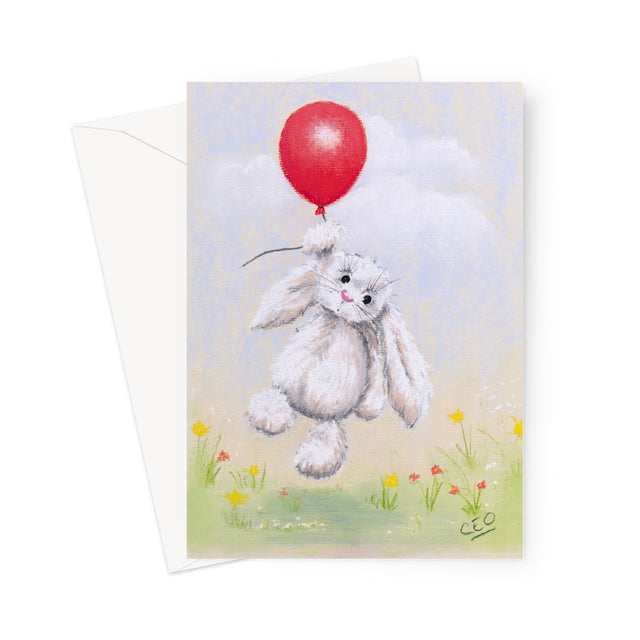 A greeting card featuring a whimsical illustration of a fluffy white bunny floating in the air while holding a bright red balloon, with a soft pastel sky, clouds, and small wildflowers below. A white envelope is partially visible behind the card on a clean white background.